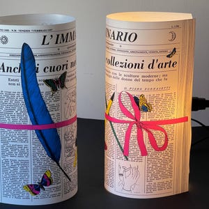 May include: Two cylindrical lamps shaped like rolled-up newspapers with colorful illustrations. The lamp on the left has a blue feather and pink ribbon, while the lamp on the right has a pink ribbon and a pencil. Both lamps have text in Italian, including the words "L'IMMA" and "collezioni d'arte".