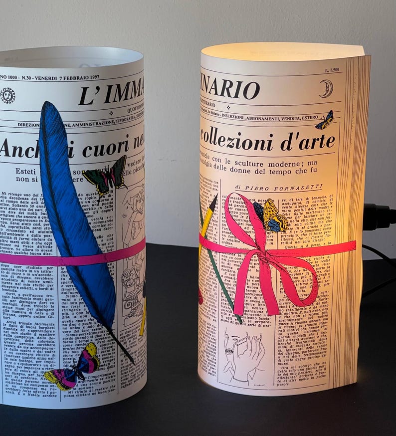 May include: Two cylindrical lamps shaped like rolled-up newspapers with colorful illustrations. The lamp on the left has a blue feather and pink ribbon, while the lamp on the right has a pink ribbon and a pencil. Both lamps have text in Italian, including the words "L'IMMA" and "collezioni d'arte".