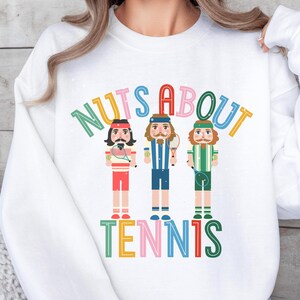 May include: White sweatshirt with a colorful graphic of three nutcracker figures playing tennis. The text "NUTS ABOUT TENNIS" is written in a colorful arc above the figures.