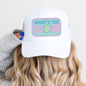 May include: White trucker hat with a mesh back and a rectangular patch. The patch features the text "WHAT'S THE SCORE" in pink and blue, with a tennis ball graphic.