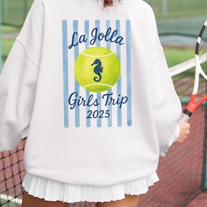 May include: White sweatshirt with blue vertical stripes and the text "La Jolla Girls Trip 2025". A yellow tennis ball with a seahorse graphic is in the center. The person is holding a tennis racket.