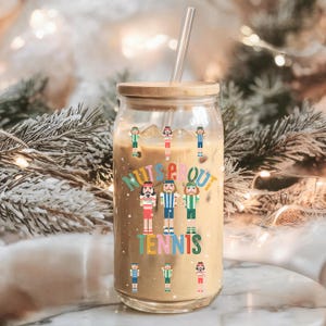 May include: A clear glass tumbler with a wooden lid and a clear straw. The tumbler is filled with a light brown beverage and ice cubes. The words "NUTS ABOUT TENNIS" are printed on the glass, along with cartoon tennis player figures.