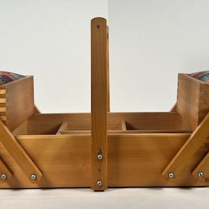 May include: A wooden sewing box with a handle and two hinged compartments. Each compartment has a colorful patchwork cushion. The box is made of light-colored wood and has a classic design, perfect for storing sewing supplies.