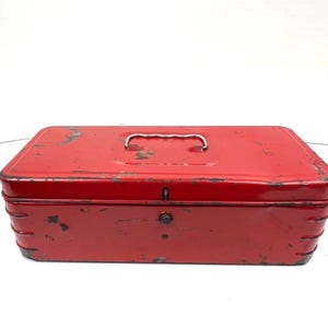 May include: A rectangular red metal tool box featuring a hinged lid, a silver-toned metal handle on top, and a central latch mechanism. The exterior surface shows signs of wear with visible paint chips and scratches along the edges and corners.