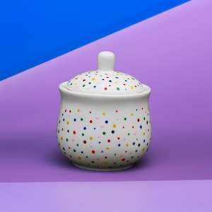 Op de afbeelding: Een witte keramische suikerpot met deksel, versierd met kleurrijke stippen in rood, geel, groen en blauw. De pot heeft een ronde vorm en een klein handvat op het deksel. De achtergrond is een verloop van blauw en paars.