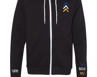 May include: Black zip-up hoodie with a white zipper and white drawstrings. The hoodie has a small embroidered design on the left chest of three upward-facing arrows in blue, white, and yellow. The left sleeve has the text "name" embroidered in white. The right sleeve has the text "the lucky few" embroidered in blue, white, and yellow.