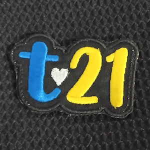 May include: A black felt patch with the letters "t21" in yellow and blue thread. A small white heart is between the "t" and the "21".
