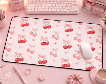 Può includere: Un tappetino per mouse rettangolare rosa con bordo nero, con un motivo di ciliegie rosse legate con fiocchi rosa. Un mouse per computer bianco è appoggiato sul tappetino. Lo sfondo include una tastiera, accessori da scrivania e una tazza di caffè.
