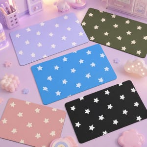 May include: A collection of rectangular desk mats in various colors, including lavender, blue, pink, black, and olive green. Each mat features a repeating pattern of white stars. The mats are arranged on a light purple surface.