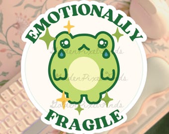 Op de afbeelding: Een ronde sticker met een groene cartoonkikker en de woorden "Emotionally Fragile" in het groen. De kikker heeft grote ogen, tranen en glitters. De sticker ligt op een lichtroze ondergrond.