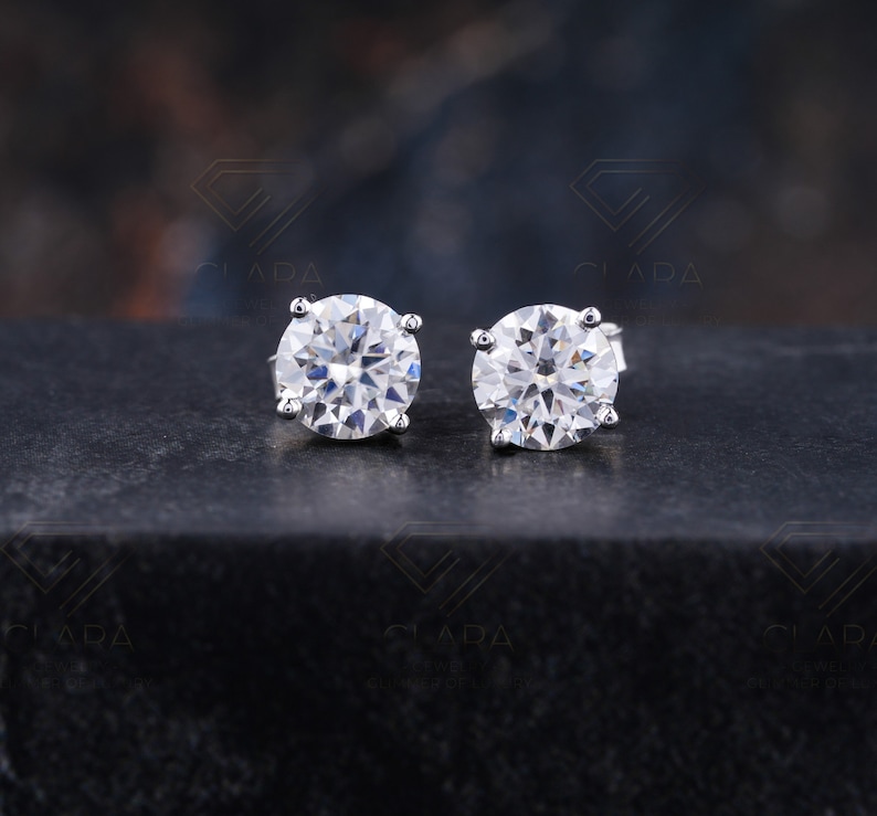 May include: A pair of round, clear gemstone stud earrings. Each earring features a large, faceted stone set in a silver or white metal prong setting. The earrings are displayed on a dark, textured surface.