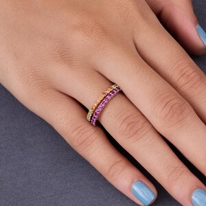 May include: Two delicate rings with colorful gemstones are worn on a finger. One ring features a gradient of orange to yellow stones, while the other showcases pink and purple gems. The rings are set against a dark gray background.