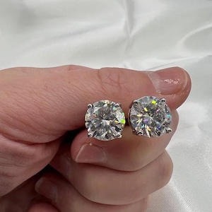 May include: A pair of sparkling, round-cut diamond stud earrings. The diamonds are held in a four-prong setting, reflecting light and showcasing their brilliance. The earrings are held in a hand against a white background.