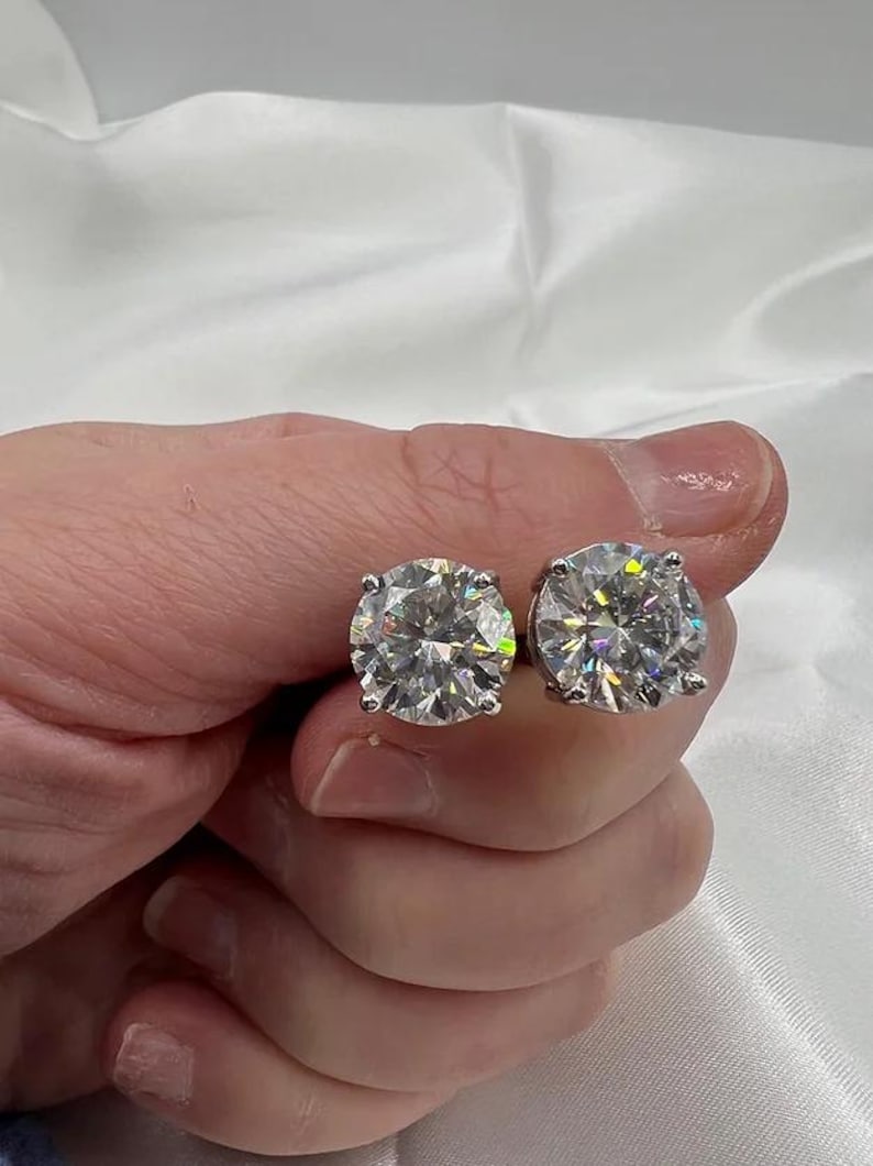May include: A pair of sparkling, round-cut diamond stud earrings. The diamonds are held in a four-prong setting, reflecting light and showcasing their brilliance. The earrings are held in a hand against a white background.