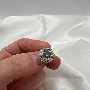 May include: A round, brilliant-cut diamond with multiple facets, reflecting light. The colorless gemstone is held up against a white satin background. The diamond is a classic piece of jewelry.