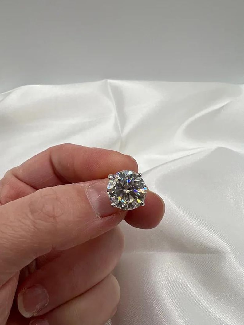 May include: A round, brilliant-cut diamond with multiple facets, reflecting light. The colorless gemstone is held up against a white satin background. The diamond is a classic piece of jewelry.