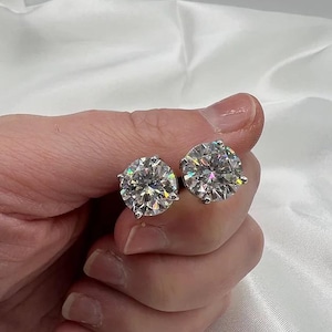 Puede incluir: Un par de brillantes pendientes de diamantes redondos. Los pendientes se sostienen en una mano, mostrando su brillo sobre un fondo blanco y suave. Los pendientes están engastados en un metal plateado.