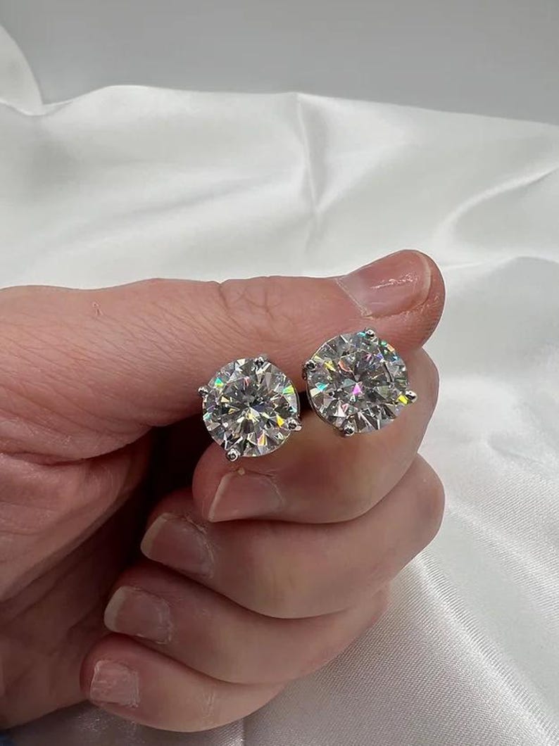May include: A pair of sparkling, round-cut diamond stud earrings. The earrings are held in a hand, showcasing their brilliance against a soft, white background. The earrings are set in a silver metal.
