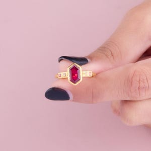 May include: A gold ring featuring a large, faceted, red gemstone in a hexagonal shape. The ring has a delicate band with small, rectangular accents on either side of the gemstone. The ring is held against a soft pink background.