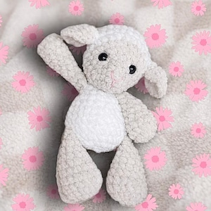 May include: A handmade crochet lamb toy in shades of white and grey, with black eyes and a pink nose. The lamb is posed on a white background with pink flower accents. This soft toy is perfect for a baby.