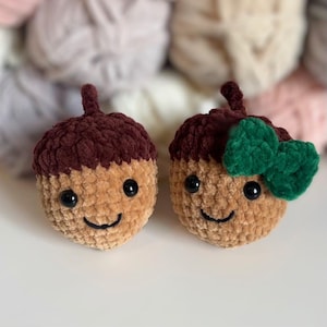 Puede incluir: Dos juguetes de peluche con forma de bellota. Las tapas de las bellotas son de color marrón oscuro y los cuerpos son de color canela. Cada uno tiene ojos negros y una sonrisa cosida. Uno tiene un lazo verde.