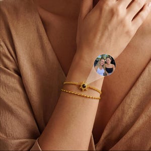 Può includere: Un bracciale dorato a doppio filo con un ciondolo centrale a forma di sole. Il ciondolo contiene una piccola proiezione circolare che mostra una fotografia di due persone. Il bracciale è composto da una catena testurizzata e una catena a perline.
