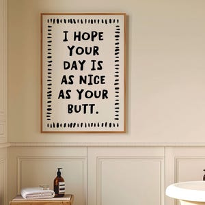 May include: A bathroom scene featuring a framed print with the text "I HOPE YOUR DAY IS AS NICE AS YOUR BUTT." The print is in a wooden frame. A small wooden stool holds a rolled white towel and a brown bottle. The floor has a black and white checkered pattern.