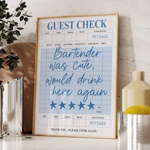 Peut inclure: Impression d'un "Guest Check" encadré, avec un fond quadrillé bleu clair et le texte "Bartender was cute, would drink here again". L'impression est dans un cadre doré, sur un comptoir blanc.