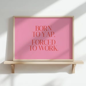 May include: A framed print with a pink background and the text "BORN TO YAP, FORCED TO WORK" in red. The print is displayed on a light wood shelf against a white wall. The frame is a light wood colour.