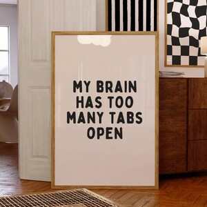 May include: A framed print with a white background and black text that reads "My brain has too many tabs open". The print is hanging on a wall in a room with a wooden floor and a wooden dresser.