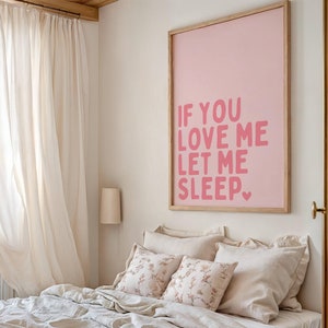 May include: A bedroom scene featuring a bed with beige bedding, pillows, and floral accent pillows. A framed pink poster with the text "IF YOU LOVE ME LET ME SLEEP" hangs above the bed. Sheer curtains frame a window.