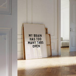 May include: A white framed poster with black text that reads "My brain has too many tabs open". The poster is leaning against a white wall.