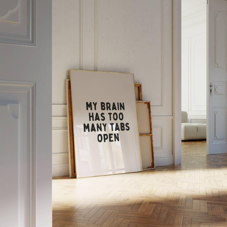 May include: A white framed poster with black text that reads "My brain has too many tabs open". The poster is leaning against a white wall.