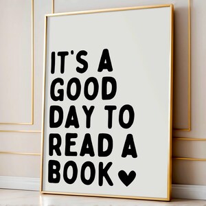 May include: A framed print with the text "IT'S A GOOD DAY TO READ A BOOK" in bold black letters on a white background. A black heart symbol is at the end of the text. The frame is gold.