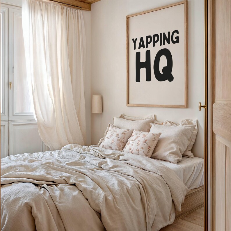 May include: A bedroom with a bed made with a beige linen duvet and pillows. The bed is next to a window with white curtains. A framed print with the text "YAPPING HQ" hangs on the wall.