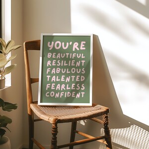 Op de afbeelding: Een wit ingelijst print met een groene achtergrond en roze tekst die "You're Beautiful Resilient Fabulous Talented Fearless Confident" zegt. De print ligt op een houten stoel.