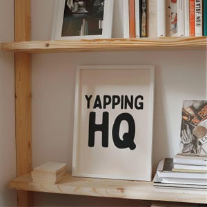 May include: A white framed print with black text that reads "YAPPING HQ" on a light pink background. The print is on a wooden shelf with other books and a framed photo.