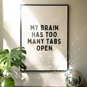 May include: A black framed print with white background and black text that reads "My brain has too many tabs open". The print is hanging on a white wall with a green plant and a silver disco ball in the foreground.