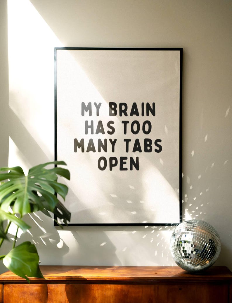 May include: A black framed print with white background and black text that reads "My brain has too many tabs open". The print is hanging on a white wall with a green plant and a silver disco ball in the foreground.