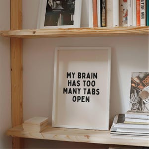 May include: A white framed print with the text "My brain has too many tabs open" in black lettering on a light pink background. The print is on a wooden shelf with other books and framed pictures.