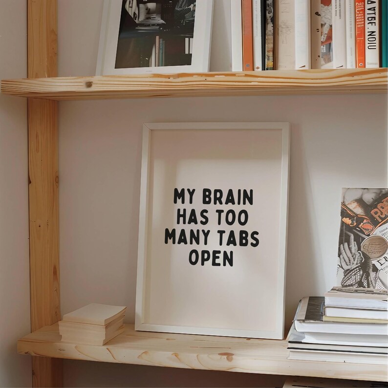 May include: A white framed print with the text "My brain has too many tabs open" in black lettering on a light pink background. The print is on a wooden shelf with other books and framed pictures.
