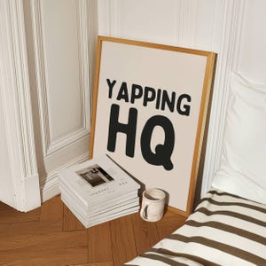 May include: A framed print with the text "YAPPING HQ" in black bold letters on a light beige background. The frame is a light wood color. The print is leaning against a stack of white journals and a white mug.