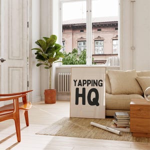 May include: A white framed poster with black text that reads "Yapping HQ" on a light wood floor in a living room with a large green plant, a couch, and a wooden side table.