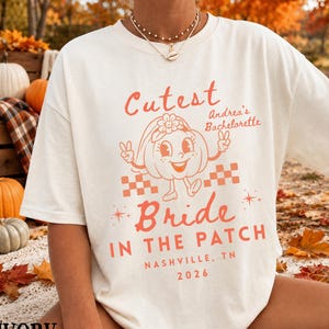 May include: An ivory short-sleeve t-shirt featuring a graphic print in orange and red. The design includes a cartoon pumpkin wearing a veil, with the text "Cutest Bride In The Patch", "Andrea's Bachelorette", "Nashville, TN", and "2026".
