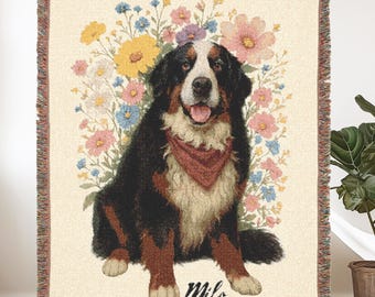 May include: Woven throw blanket featuring a Bernese Mountain Dog with a red bandana, surrounded by colorful flowers. The dog's name, "Milo," is written in script at the bottom. The blanket has a fringed edge and a cream-colored background.