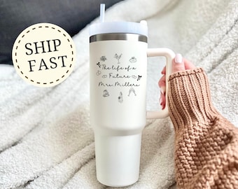 May include: A white insulated tumbler with a handle and a white lid. The tumbler has the text "The life of a Future Mrs. Millers" with wedding-themed illustrations. The tumbler is held by a person wearing a brown sweater.