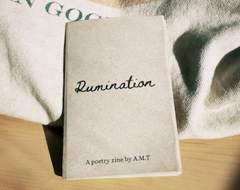 May include: A small, light-colored book titled "Rumination" with the subtitle "A poetry zine by A.M.T." The book is resting on a wooden surface, with a soft, patterned fabric in the background.