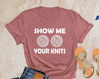 May include: A heather mauve t-shirt with the text "Show Me Your Knits" and a graphic of two yarn balls. The shirt is laid out on a light brown surface with a pair of gold sandals and blue jeans.