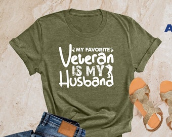 May include: A heather olive t-shirt with the text "My Favorite Veteran Is My Husband" in white. The shirt is laid flat on a textured surface with a pair of sandals and blue jeans.