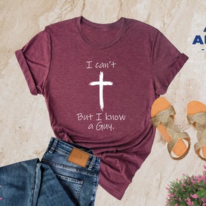 May include: A heather maroon t-shirt with the text "I can't" above a white cross and the text "But I know a Guy."  The shirt is displayed on a light brown surface with a pair of gold sandals and a pair of blue jeans.